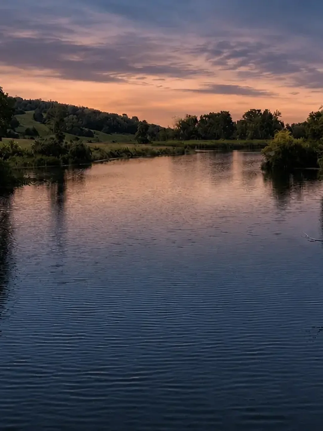 Still lake water reflecting a sunset sky between wooded banks