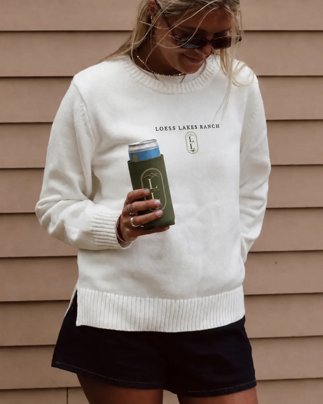 A woman wearing a cream knit crew with the LOESS LAKES RANCH wordmark and LL crest at the chest, holding an olive-green ranch koozie