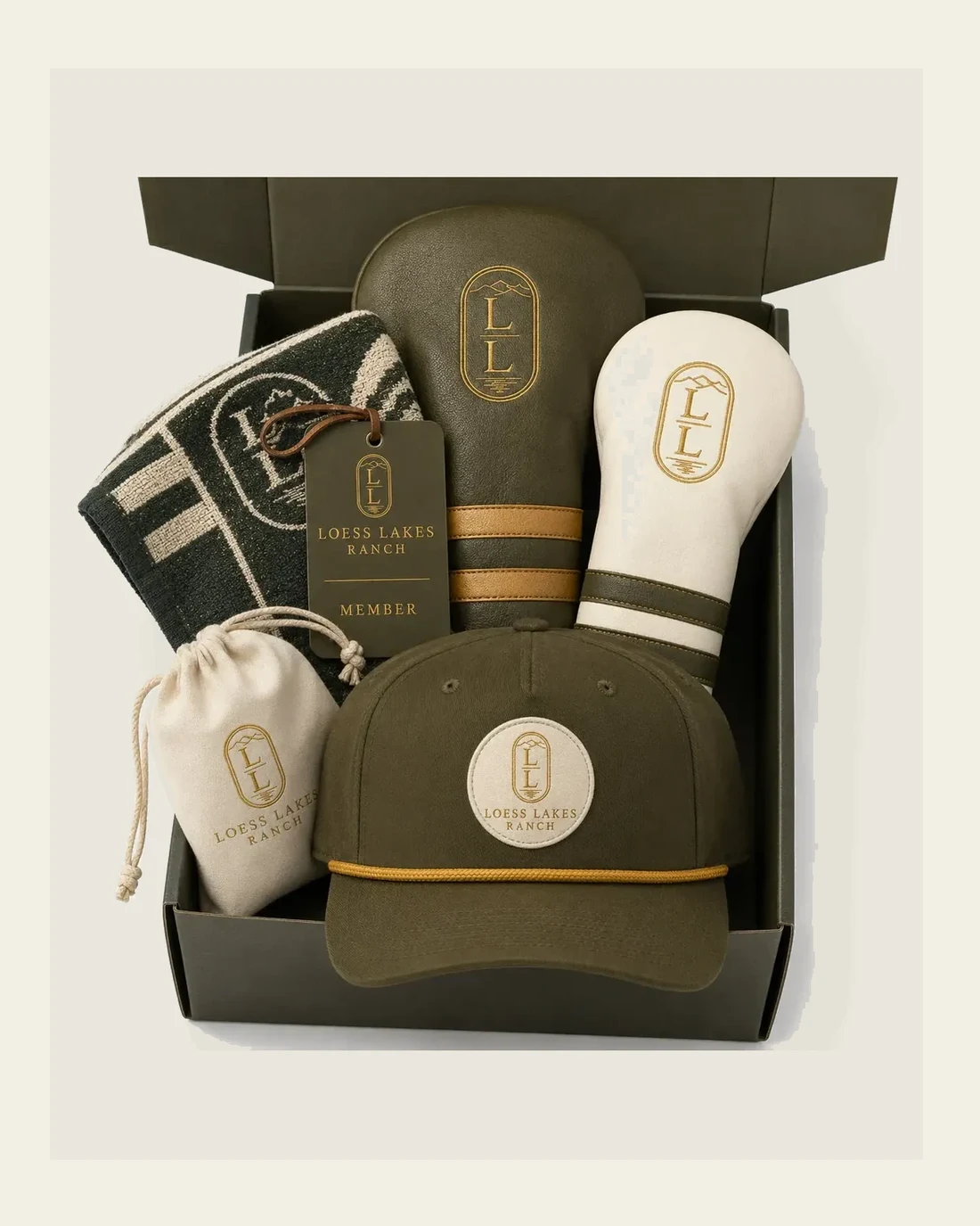 Overhead view of an olive gift box holding a rope cap, two golf headcovers, a woven towel, a member tag and a cream drawstring pouch