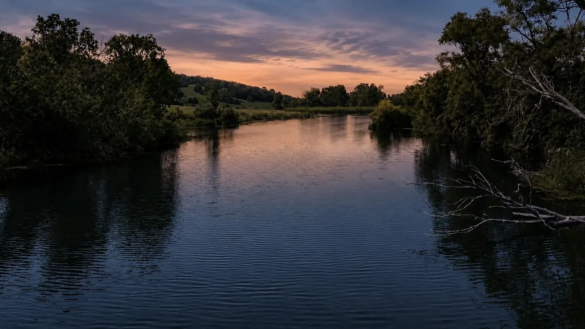 Still lake water reflecting a sunset sky between wooded banks