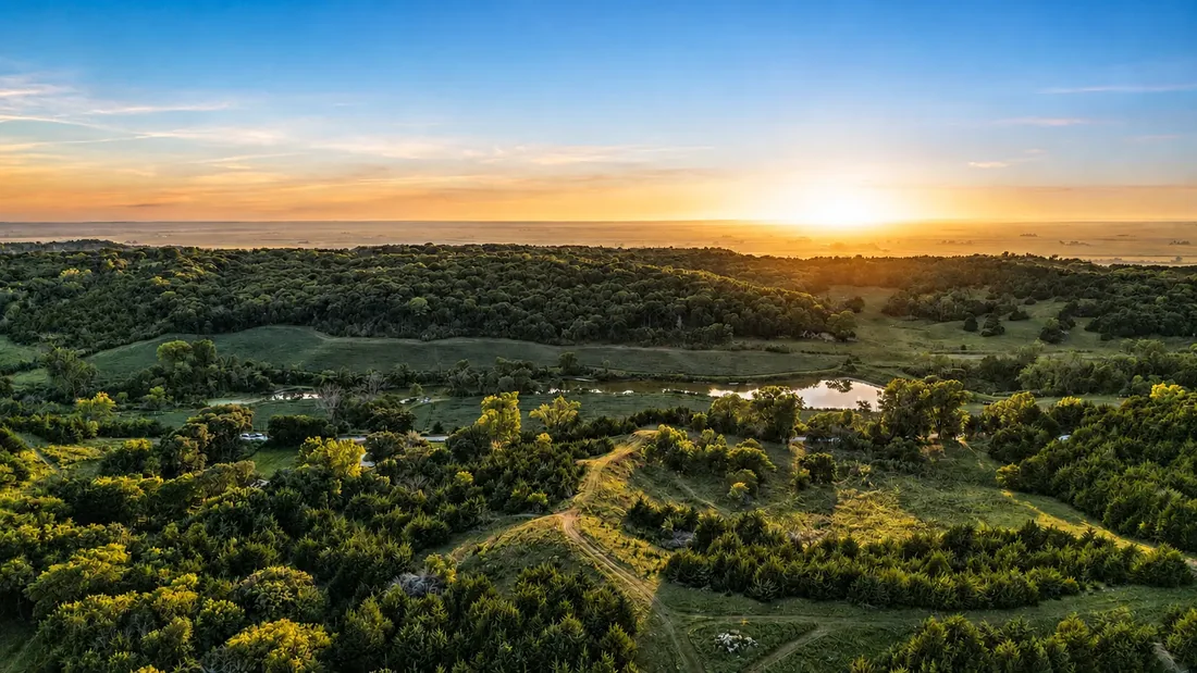 Aerial view of Loess Lakes Ranch at sunset: golden light across wooded ridges, rolling green hills and the private lake
