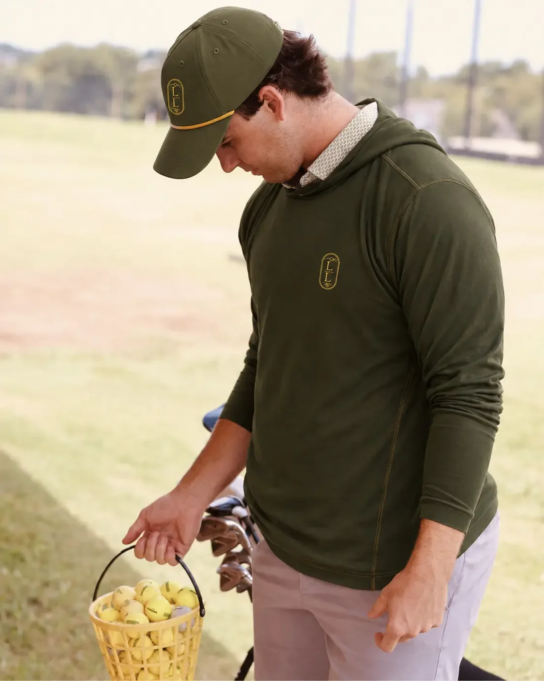 A golfer in an olive hooded performance layer and matching olive LOESS LAKES RANCH rope cap, carrying a basket of range balls