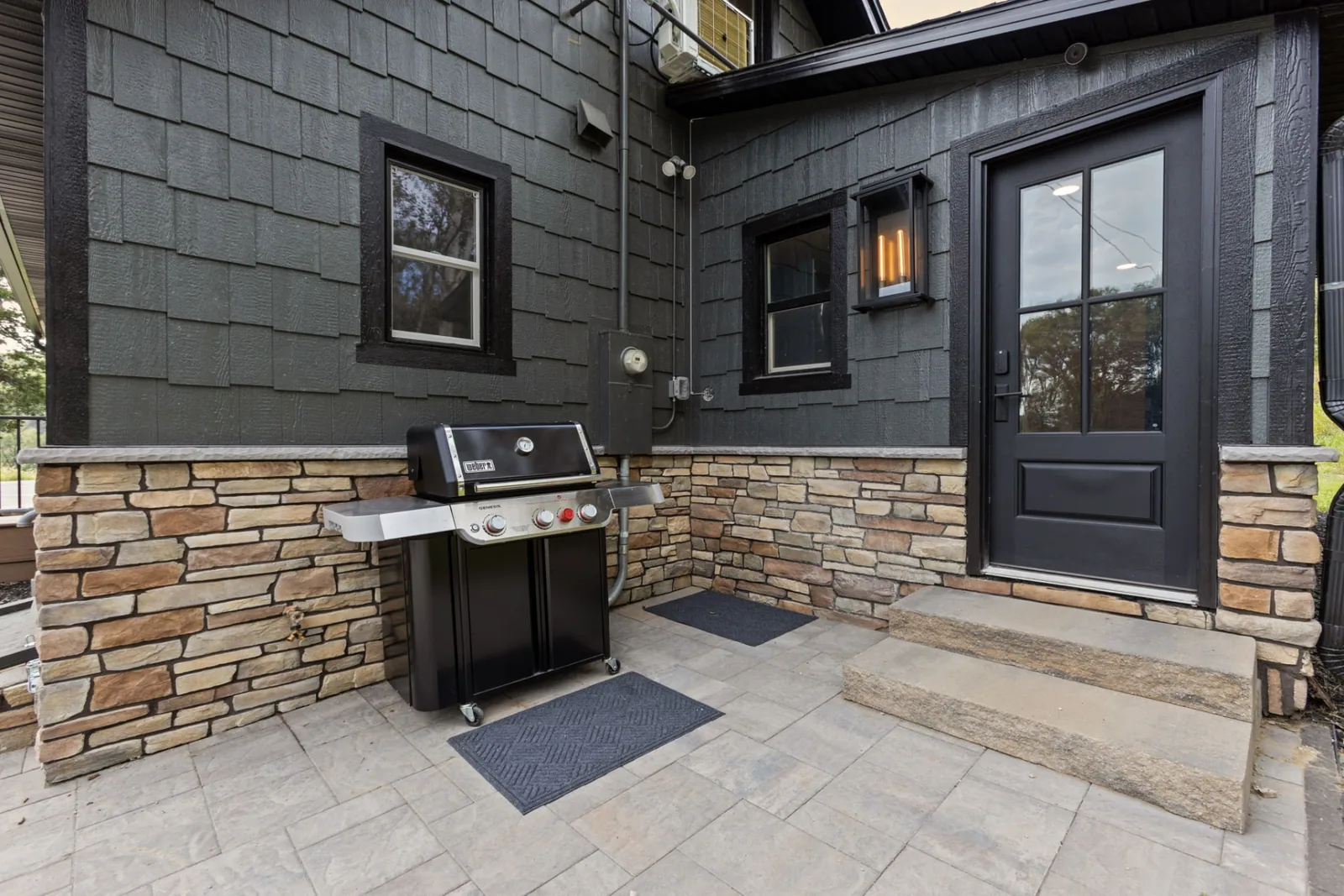 Paved patio beside the house with a gas grill, stone-clad walls, lantern sconce and the back entry door