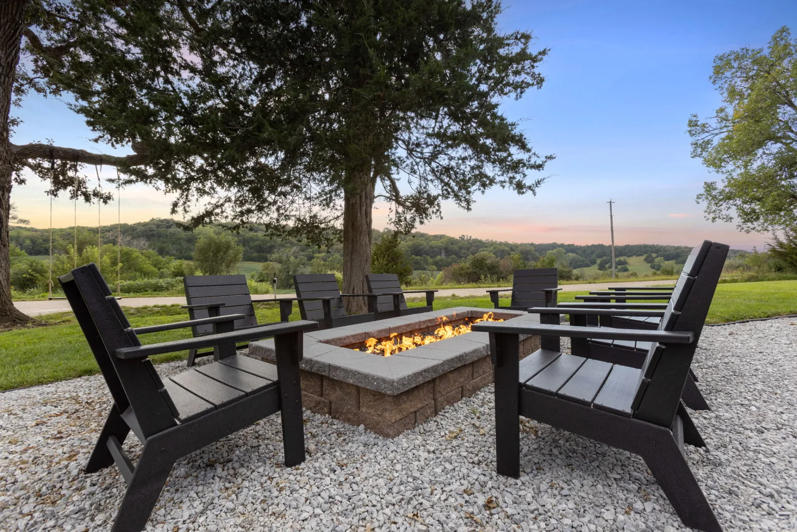 Gas firepit surrounded by black Adirondack chairs on gravel beneath a cedar tree, looking out over the rolling Loess Hills at sunset