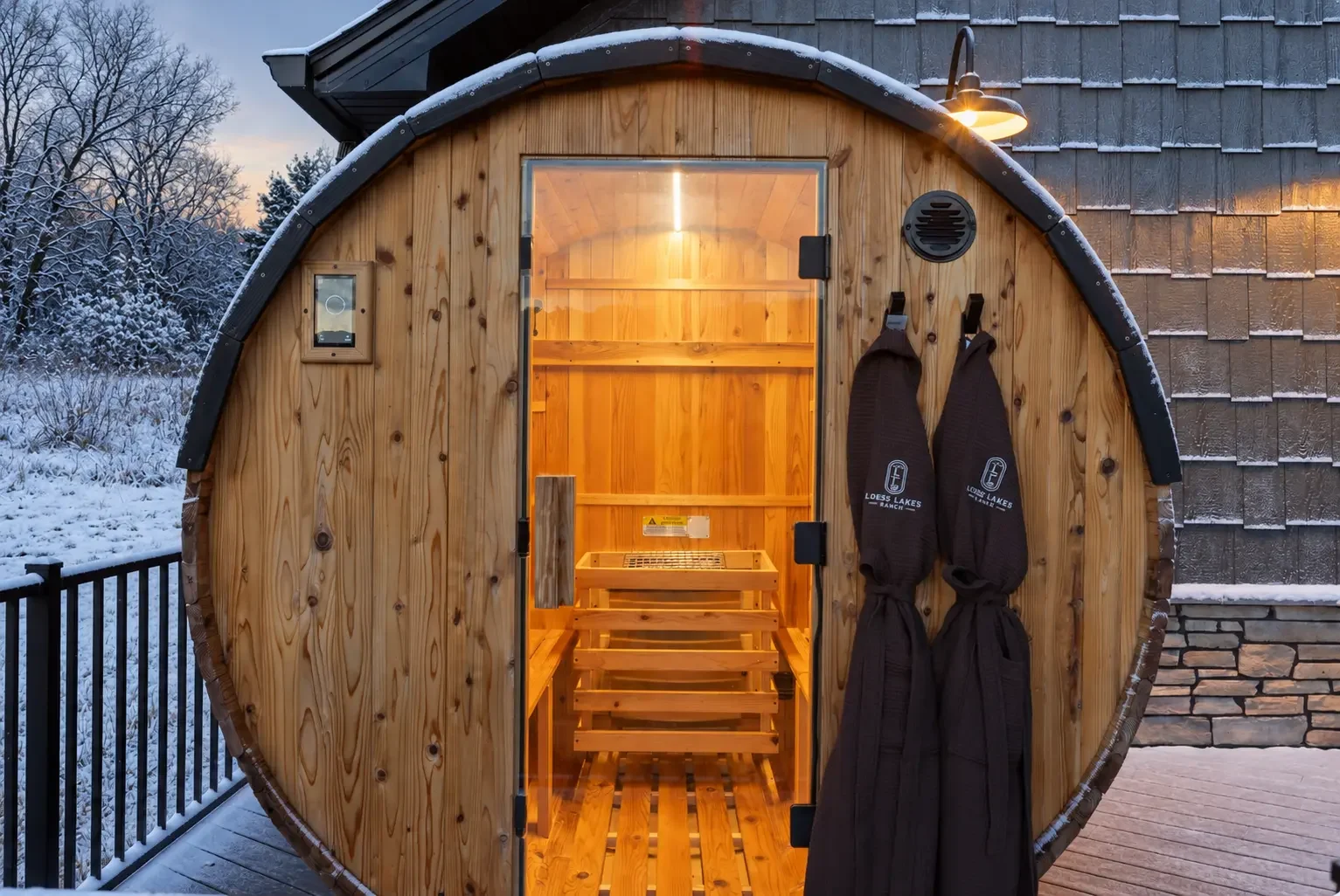 Loess Lakes Ranch barrel sauna lit from within, with two ranch robes hanging beside the door on a snowy evening
