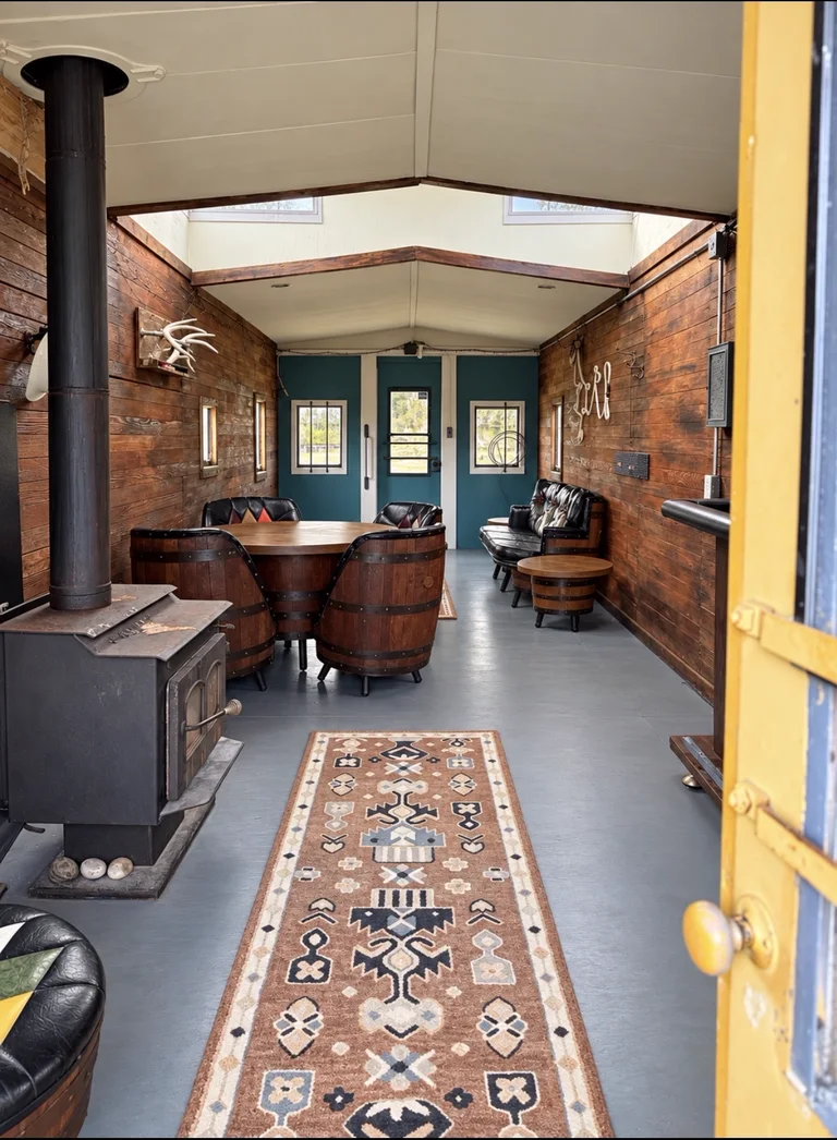 View through the open yellow caboose door into the finished interior with a barrel table, barrel chairs, leather seating and a long patterned runner