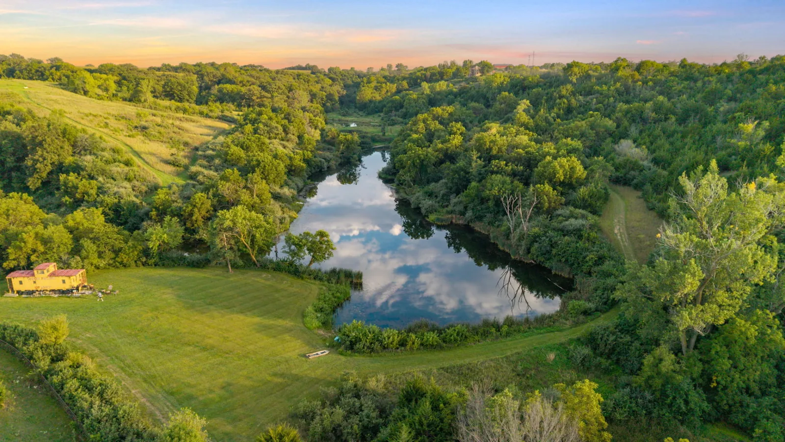 Aerial view of the private lake reflecting clouds, ringed by timber and mown lawn with the vintage caboose at the edge