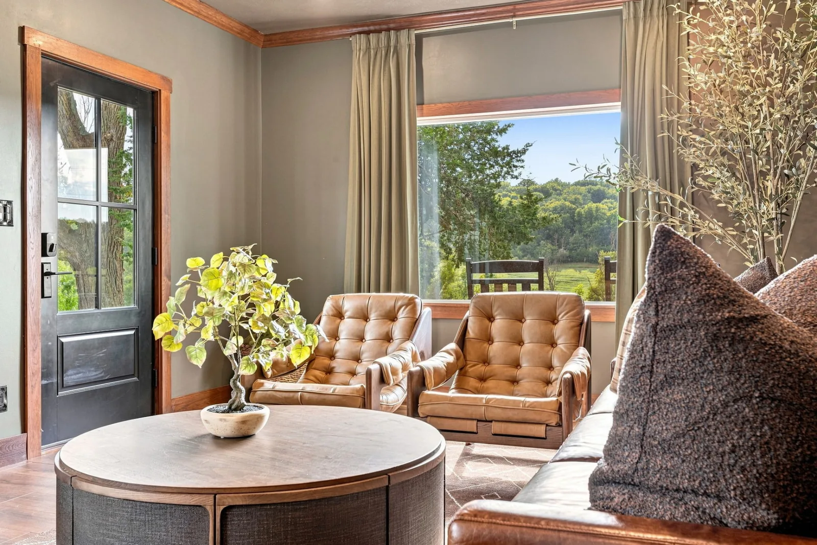 Living-room seating area with tufted leather chairs beside a large window and glass door opening to the deck and green landscape