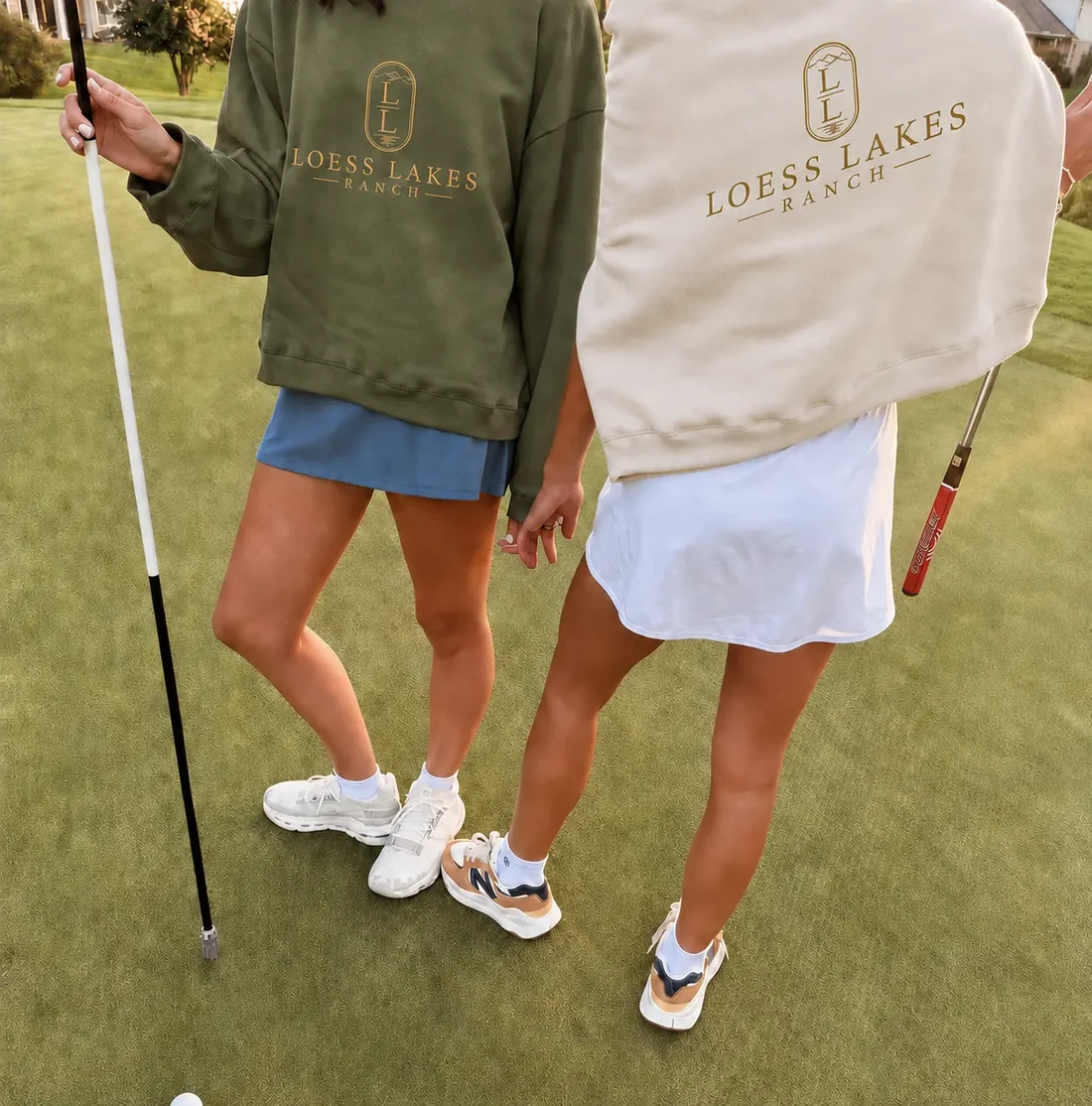 Two crewnecks on the putting green — olive and warm stone — each with the gold LOESS LAKES RANCH crest and wordmark