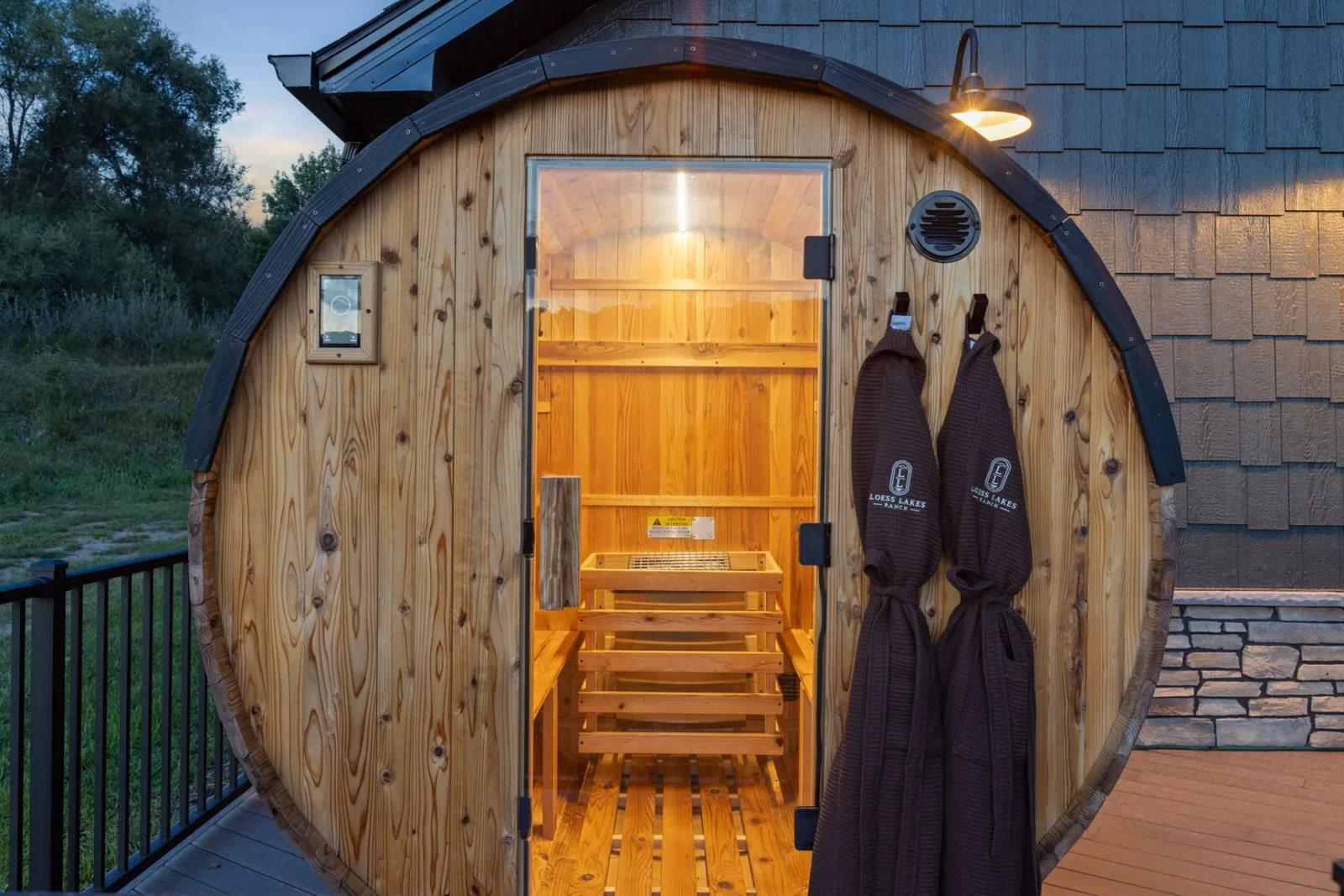 The cedar barrel sauna glowing at dusk with two Loess Lakes Ranch robes hanging beside the open glass door