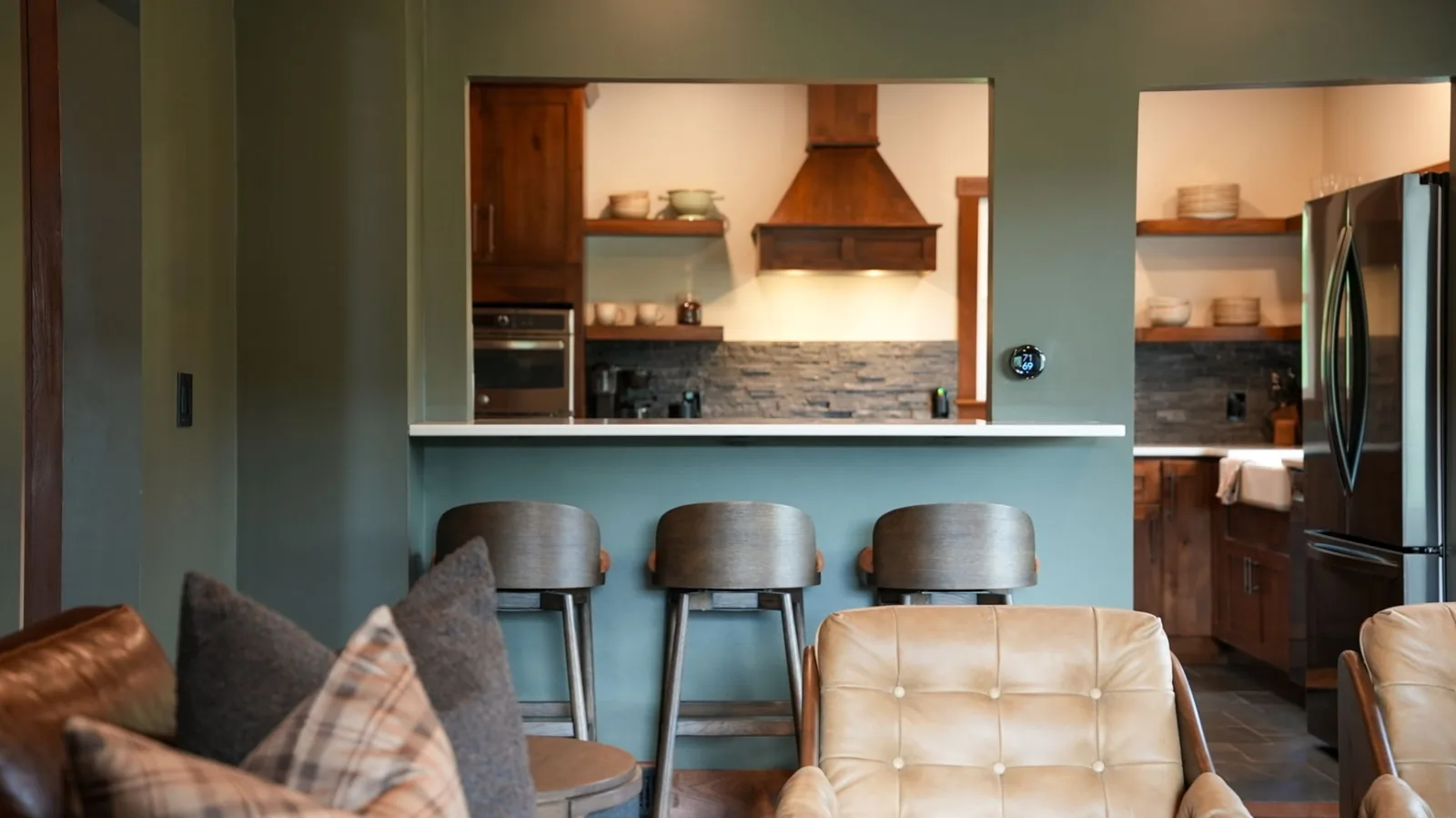 View from the living room into the kitchen, with three barstools at the breakfast bar, walnut range hood and stone backsplash