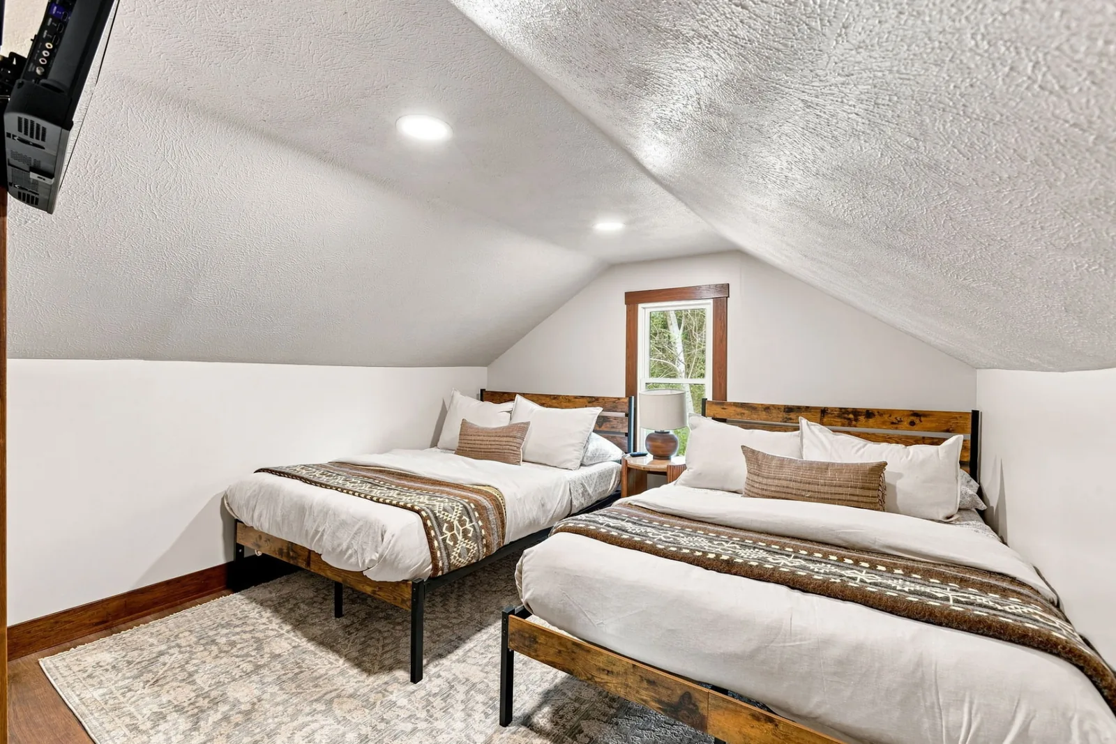 Loft bedroom under a vaulted ceiling with two beds in reclaimed-wood frames, woven throws, a patterned rug and a dormer window