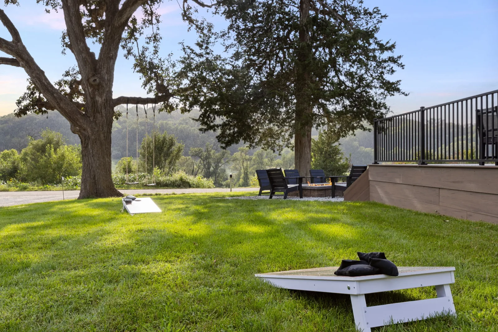 Sunlit lawn with cornhole boards, a rope tree swing under mature oaks and the gas firepit with chairs beside the deck