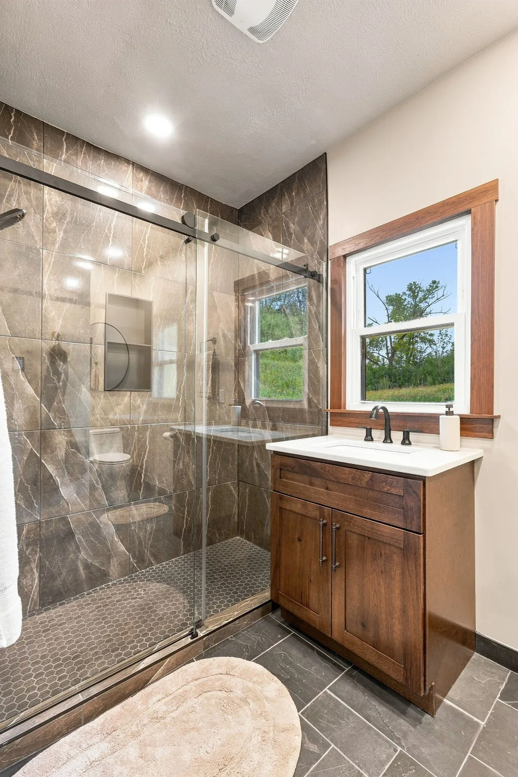 Bathroom with a large glass walk-in shower in marbled stone tile, walnut vanity and a window onto the fields