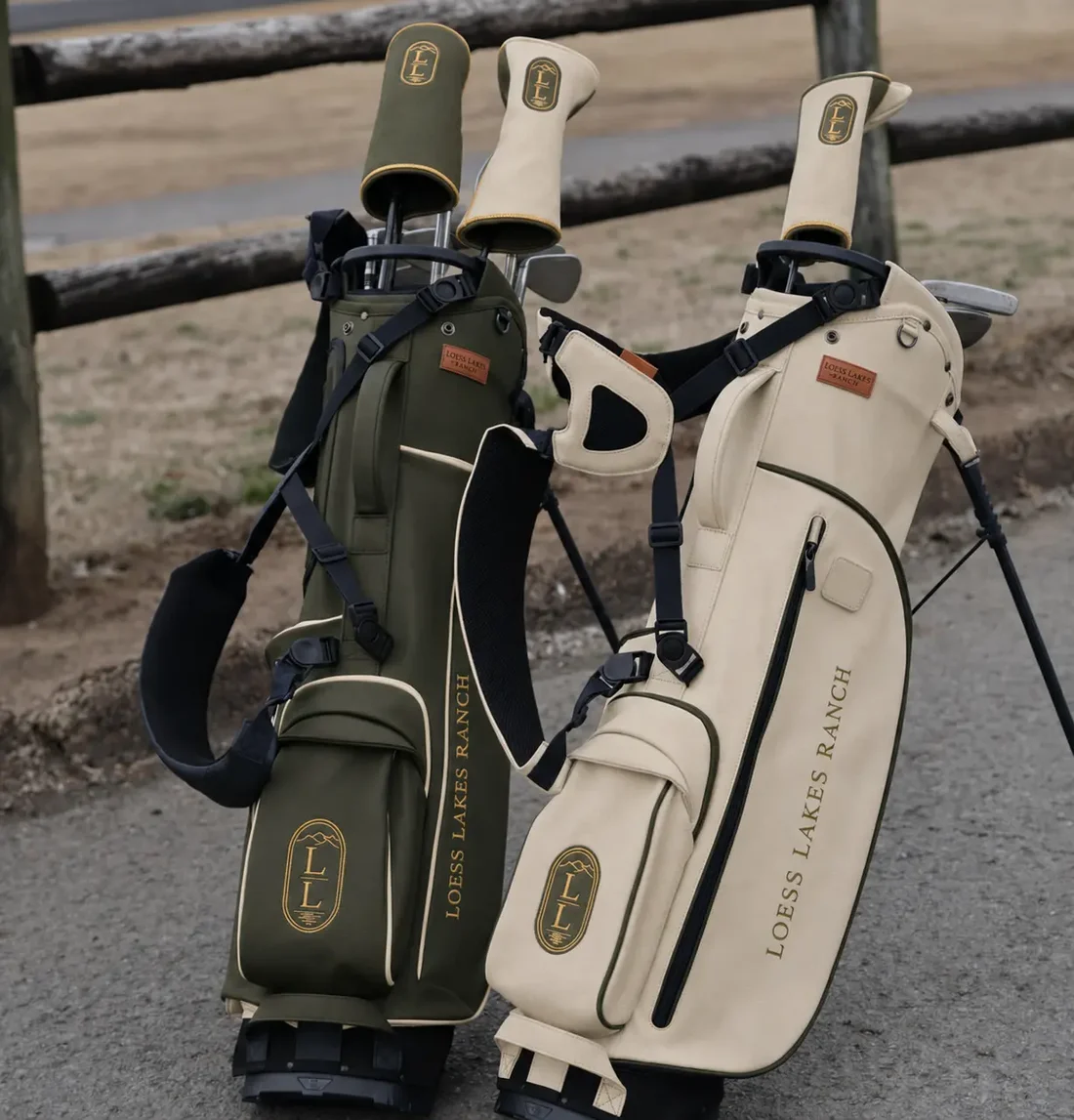 The olive and cream LOESS LAKES RANCH Heritage Stand Bags standing together against a split-rail fence with branded headcovers