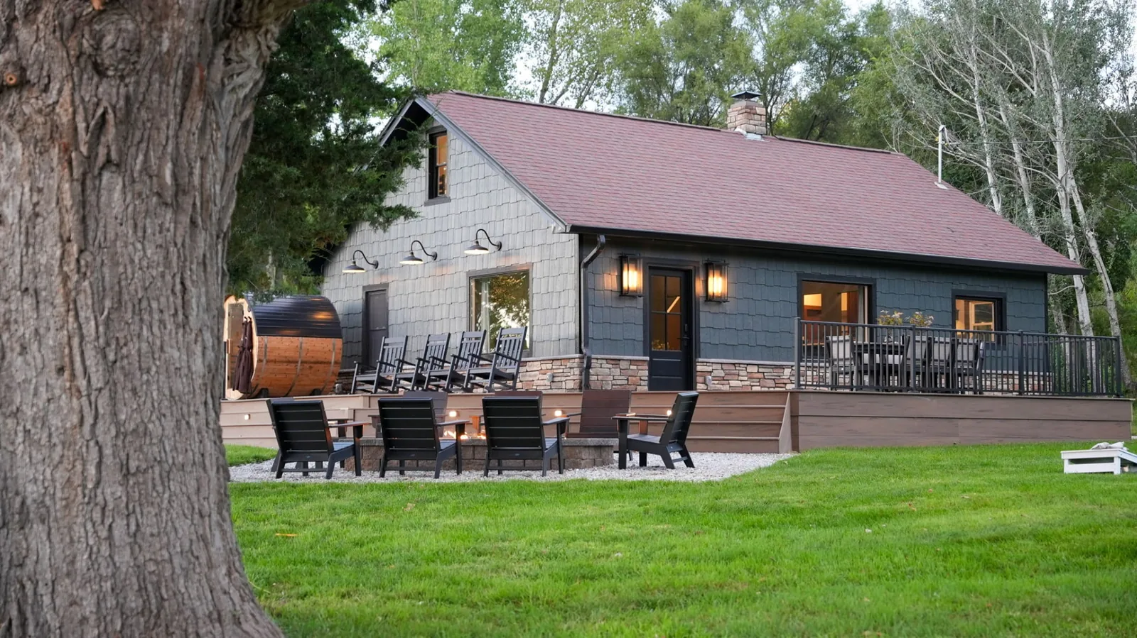 The ranch farmhouse at dusk framed by a large oak, with a lit deck, cedar barrel sauna, rocking chairs and a burning firepit ringed by black chairs on the lawn