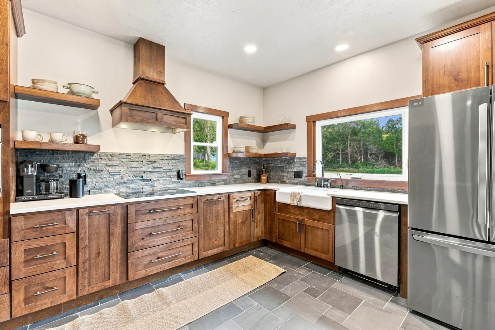 Kitchen with cherry cabinetry, quartz counters, stacked-stone backsplash, farmhouse sink and a window looking out to the trees