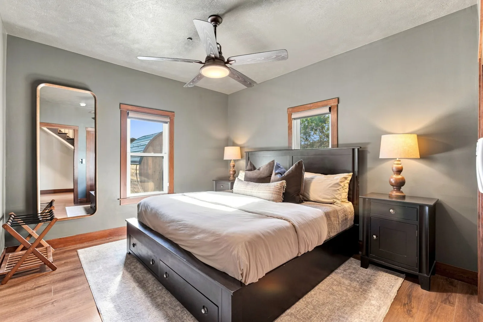 Bedroom with a black bedframe, layered linens, matching lamps, full-length mirror and a window overlooking the barrel sauna