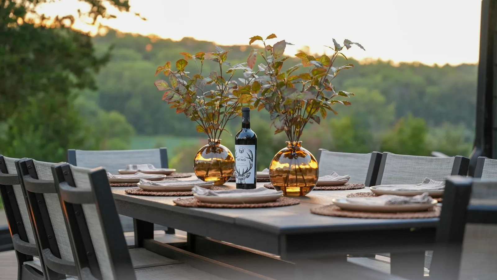 Outdoor dining table on the deck at sunset, set with plates, linen napkins, amber vases and a bottle of wine above the wooded valley